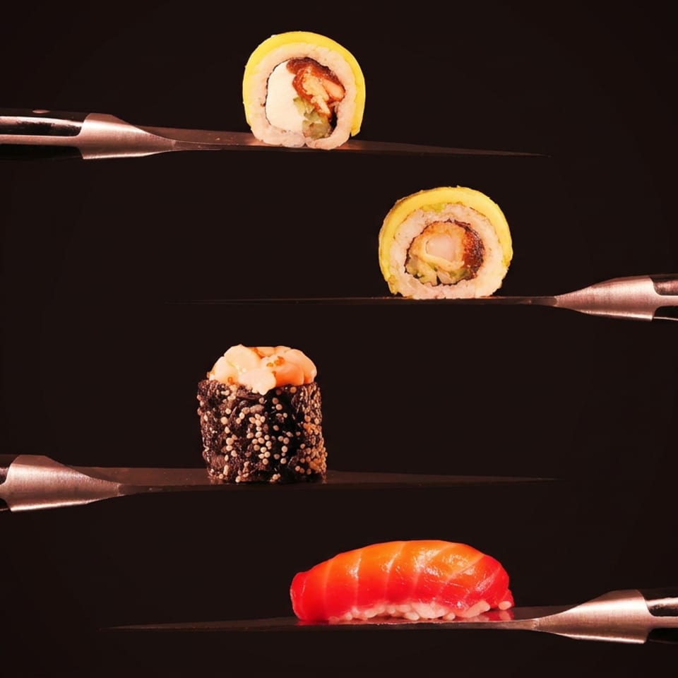 Assorted sushi pieces balanced on knives against a dark background, including rolls, a gunkan sushi, and salmon nigiri.
