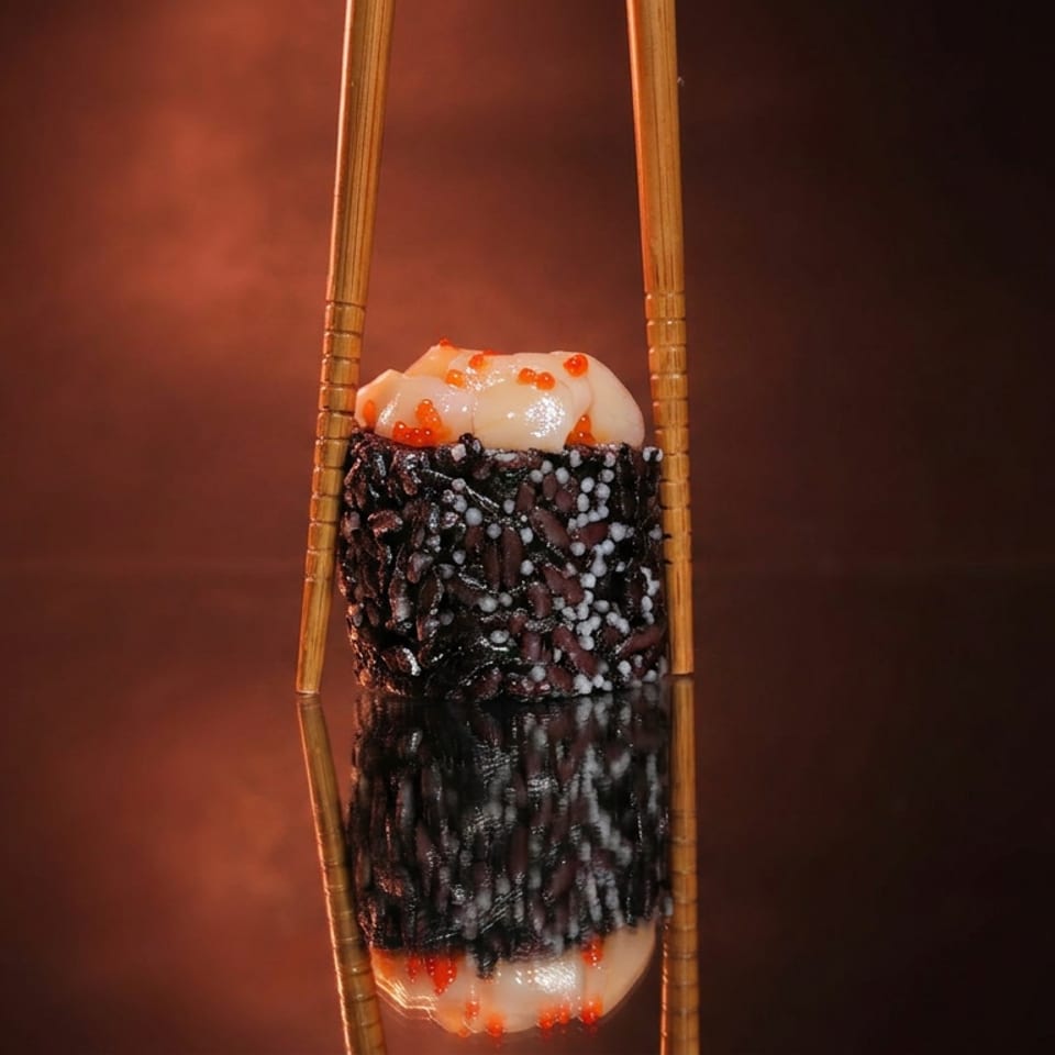 Sushi roll coated in black sesame seeds, topped with creamy sauce and orange roe, held vertically between wooden chopsticks.