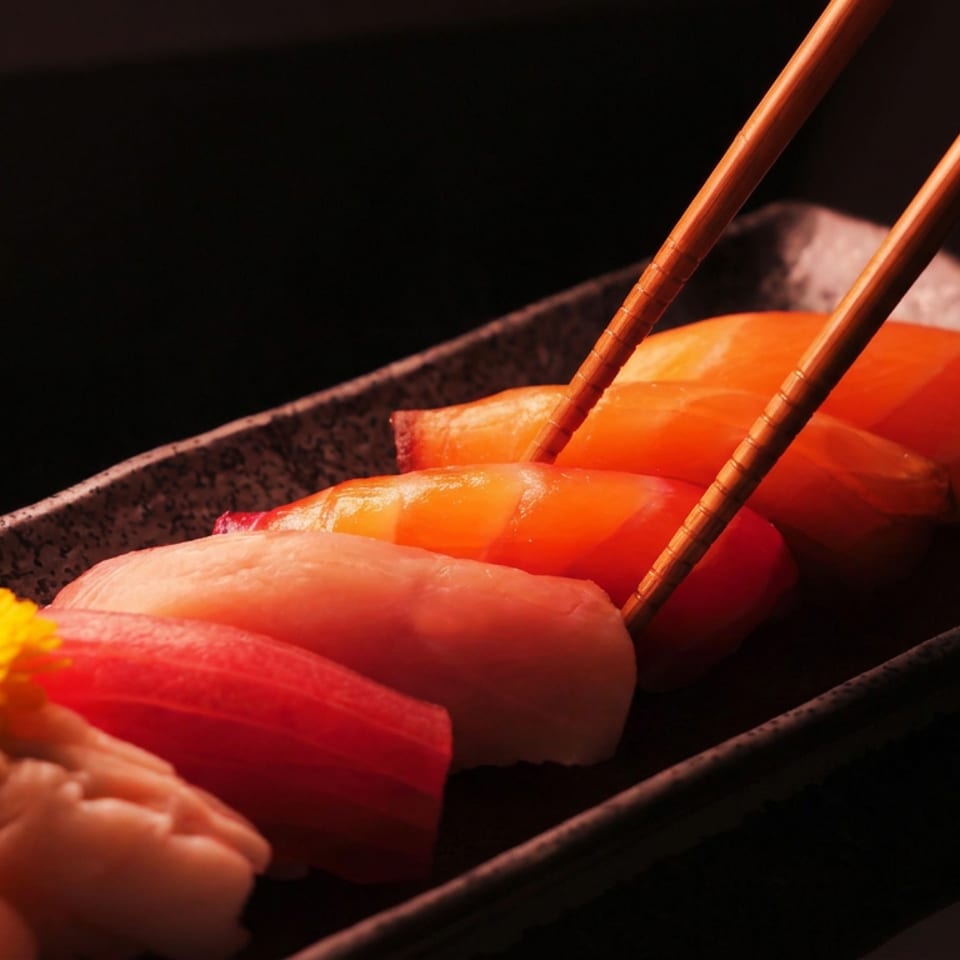 Close-up of assorted sushi nigiri on a dark rectangular plate, with chopsticks lifting one piece and a yellow flower garnish.