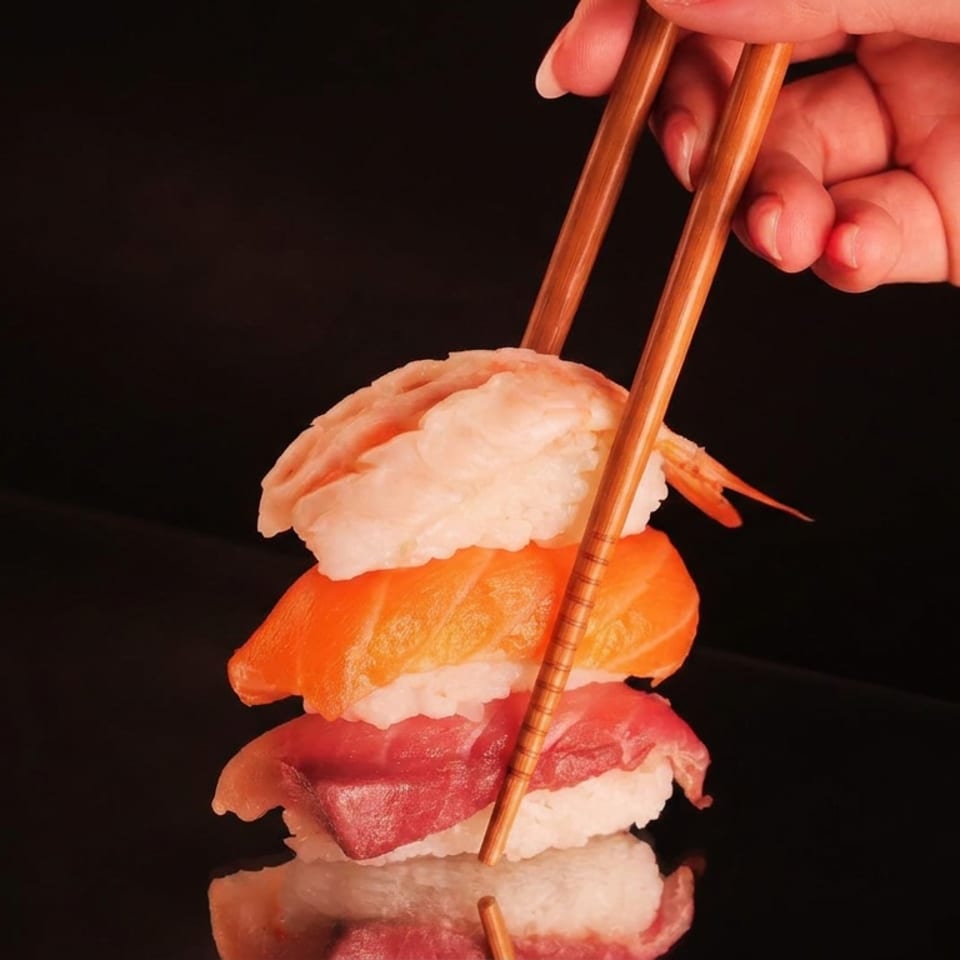 Hand using chopsticks to hold a stack of shrimp, salmon, tuna, and rice sushi against a dark background.